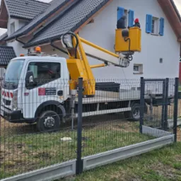 Pracownicy w podnośniku koszowym, w Tarnowskich Górach, pracują przy elewacji dwukondygnacyjnego domu z niebieskimi okiennicami.