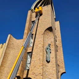 Gregtrans & Cattleya - Żółty podnośnik koszowy sięga do ceglanej wieży kościoła z krzyżem na szczycie i posągiem świętego w niszy, na tle błękitnego nieba.