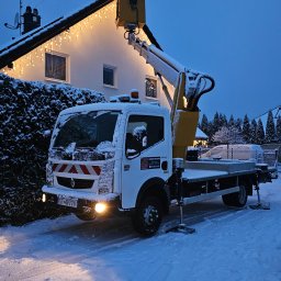 Gregtrans & Cattleya - Żółty podnośnik koszowy ustawiony na ośnieżonej posesji przed domem z dekoracyjnymi lampkami na dachu, w zimowej scenerii.