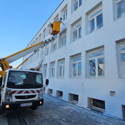 Gregtrans & Cattleya - Pracownik w podnośniku koszowym, malujący jasną elewację budynku z licznymi oknami, widoczne zabezpieczenie podłoża folią, błękitne niebo w tle.