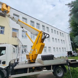 Gregtrans & Cattleya - Żółty podnośnik koszowy na białej ciężarówce z logo firmy GREGTRANS, ustawiony przy dwukolorowym budynku z wieloma oknami, drabina oparta o ścianę, widoczne ślady prac na elewacji.