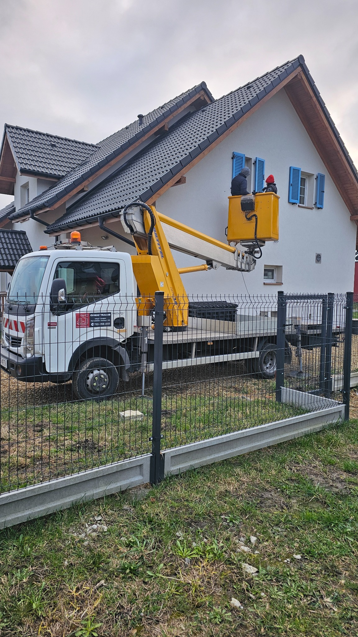 Pracownicy w podnośniku koszowym, w Tarnowskich Górach, pracują przy elewacji dwukondygnacyjnego domu z niebieskimi okiennicami.