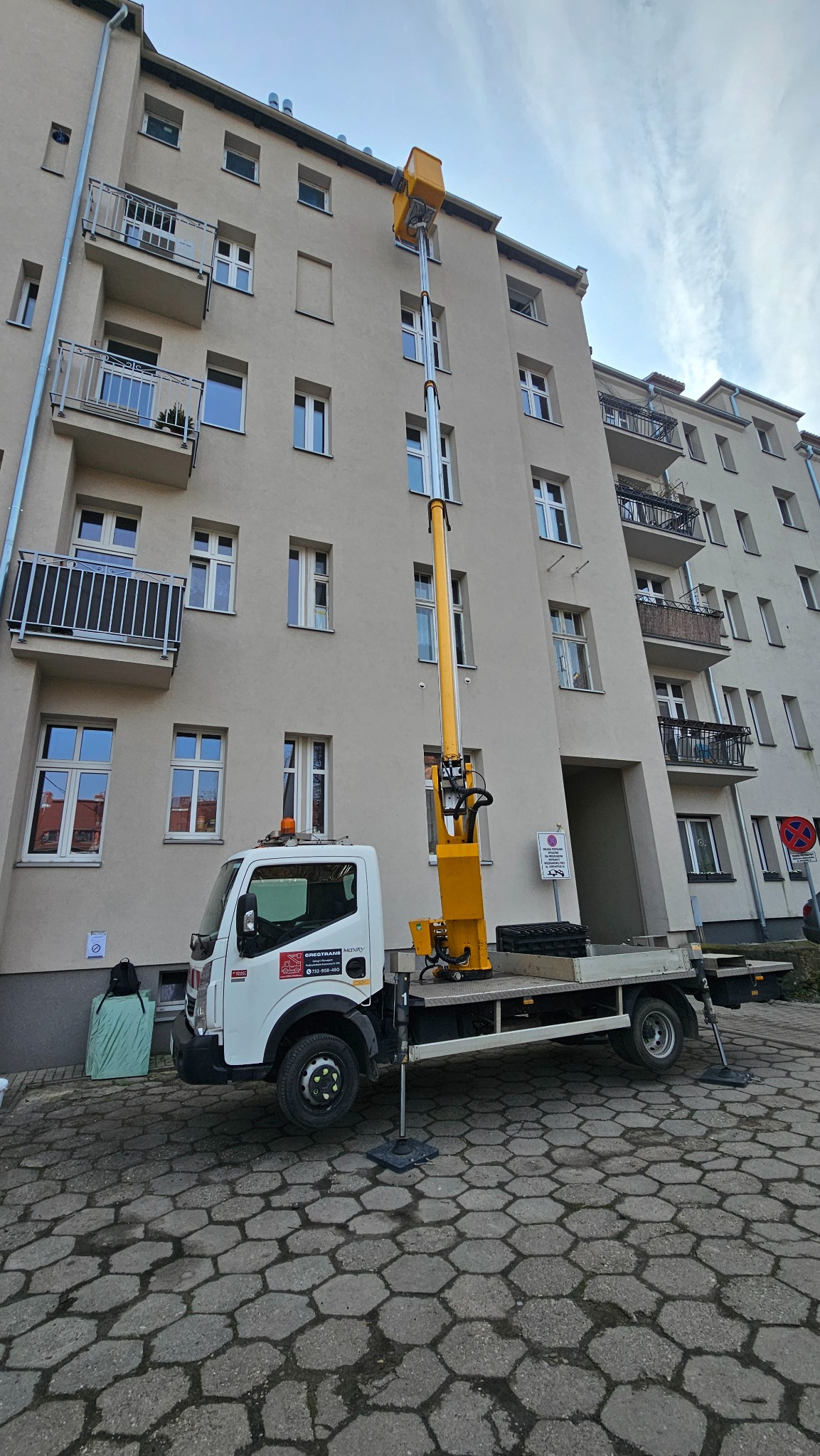 Żółty podnośnik koszowy na białej ciężarówce ustawiony przed budynkiem mieszkalnym z balkonami; widoczne podpory stabilizujące pojazd na kostce brukowej.