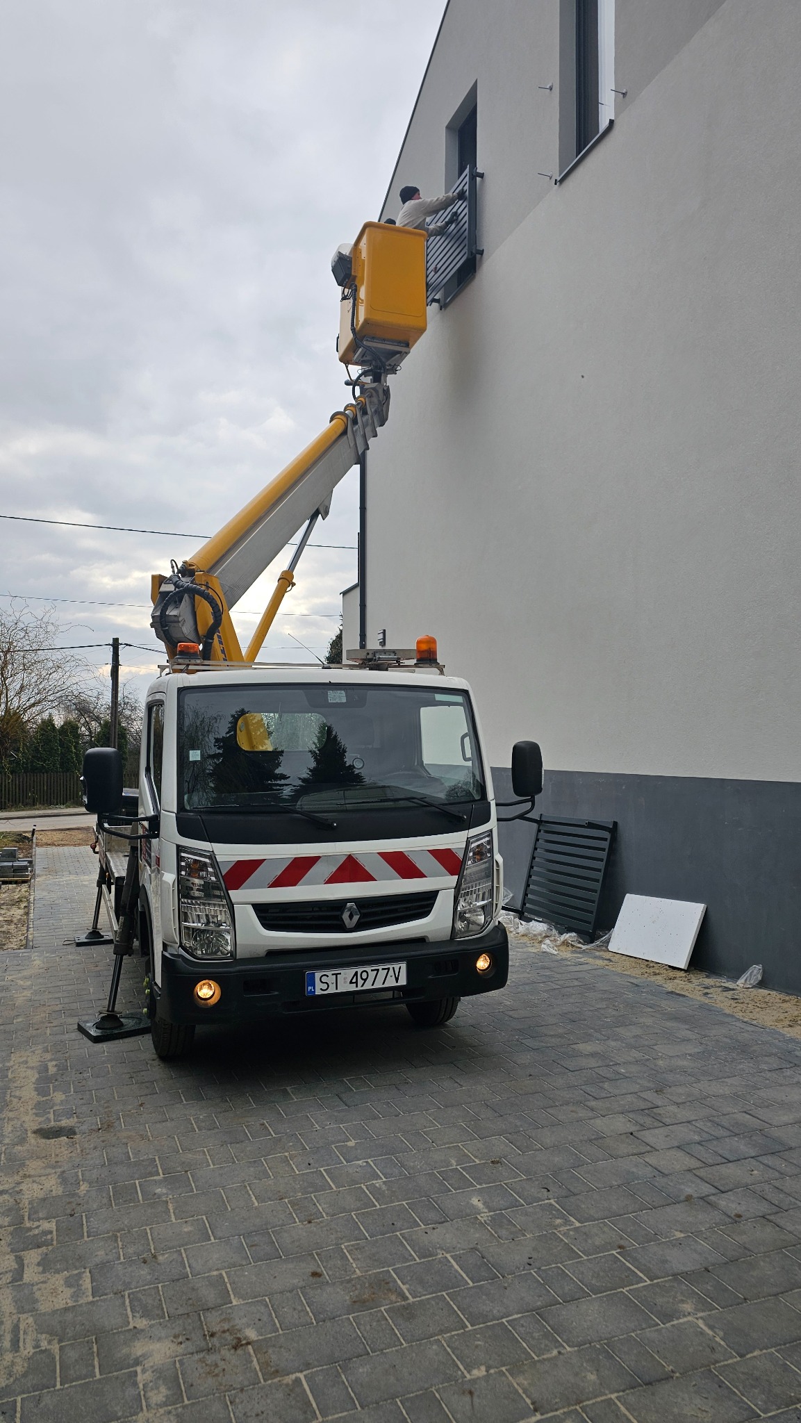 Pracownik w koszu podnośnika koszowego przy oknie na elewacji budynku, widoczny biały samochód z wysięgnikiem na tle pochmurnego nieba.