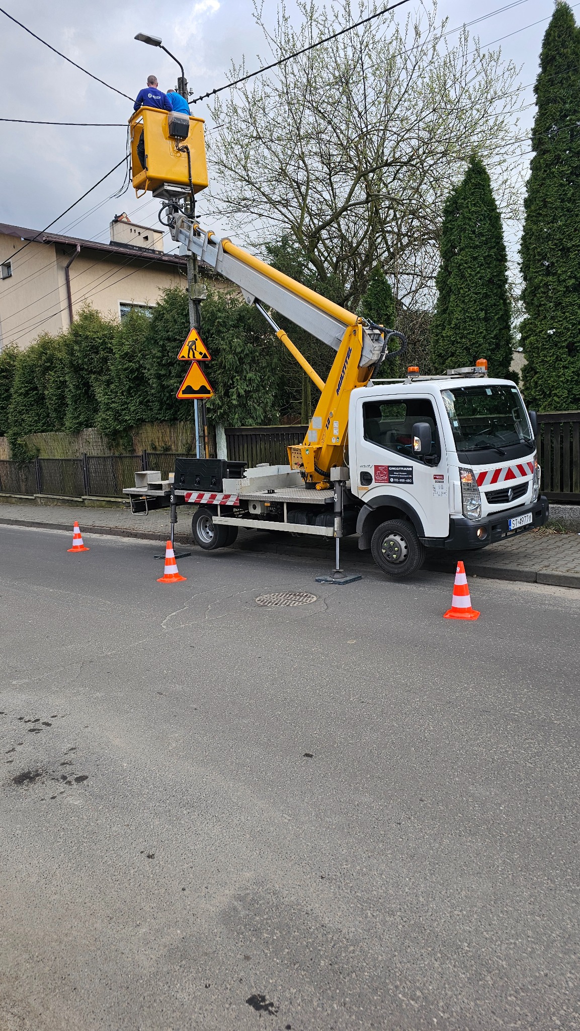 Żółty podnośnik koszowy na ulicy, pracownicy w koszu naprawiają oświetlenie uliczne, zabezpieczone pomarańczowymi pachołkami.