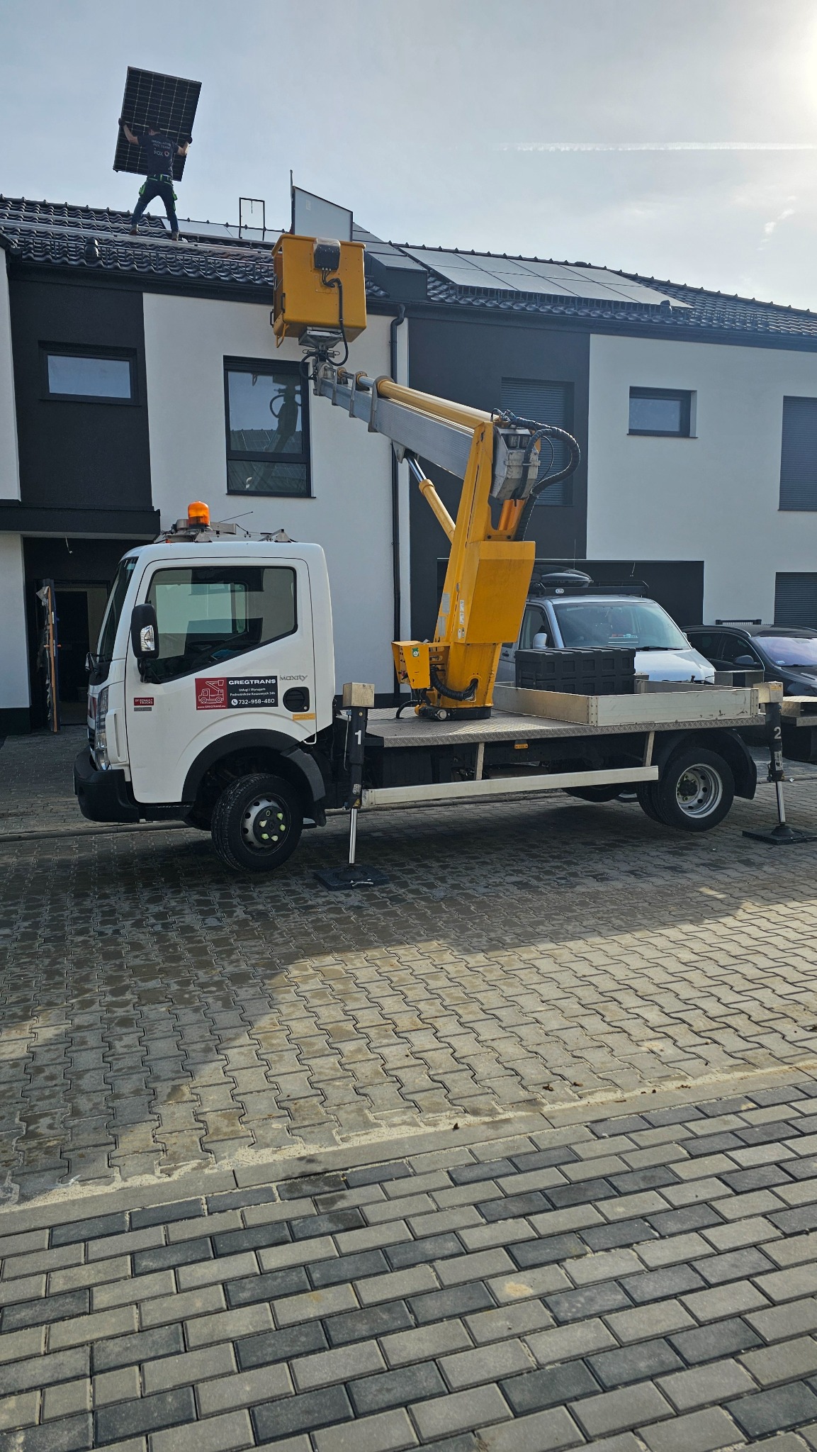 Montaż paneli słonecznych na dachu nowoczesnego domu jednorodzinnego z użyciem podnośnika koszowego, operator na dachu z panelem w rękach, widoczny fragment elewacji i brukowana nawierzchnia.