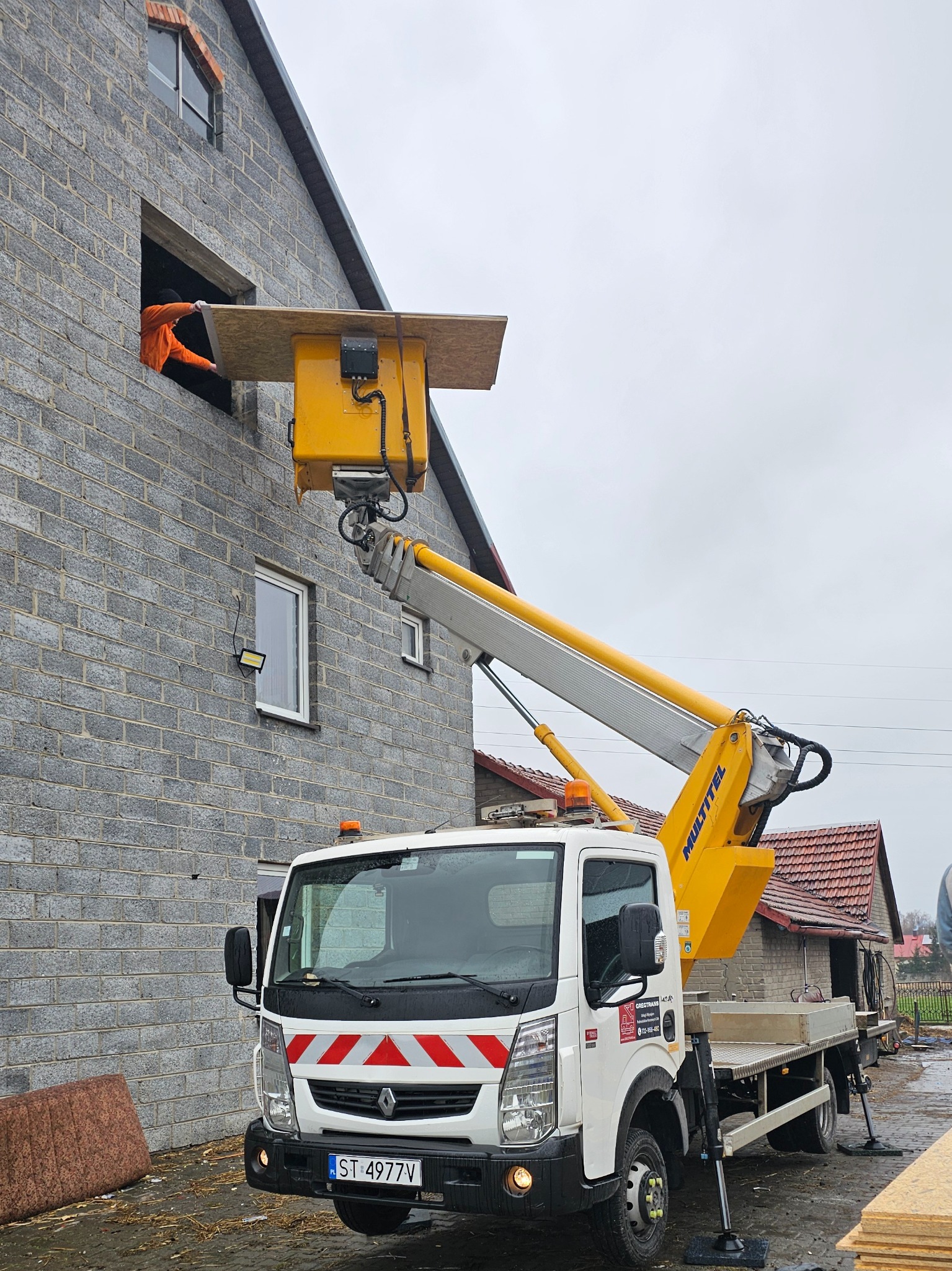 Żółto-biały podnośnik koszowy MULTITEL na tle szarego, dwupiętrowego budynku z cegły; pracownik w pomarańczowej kurtce podaje płytę z okna na drugim piętrze.