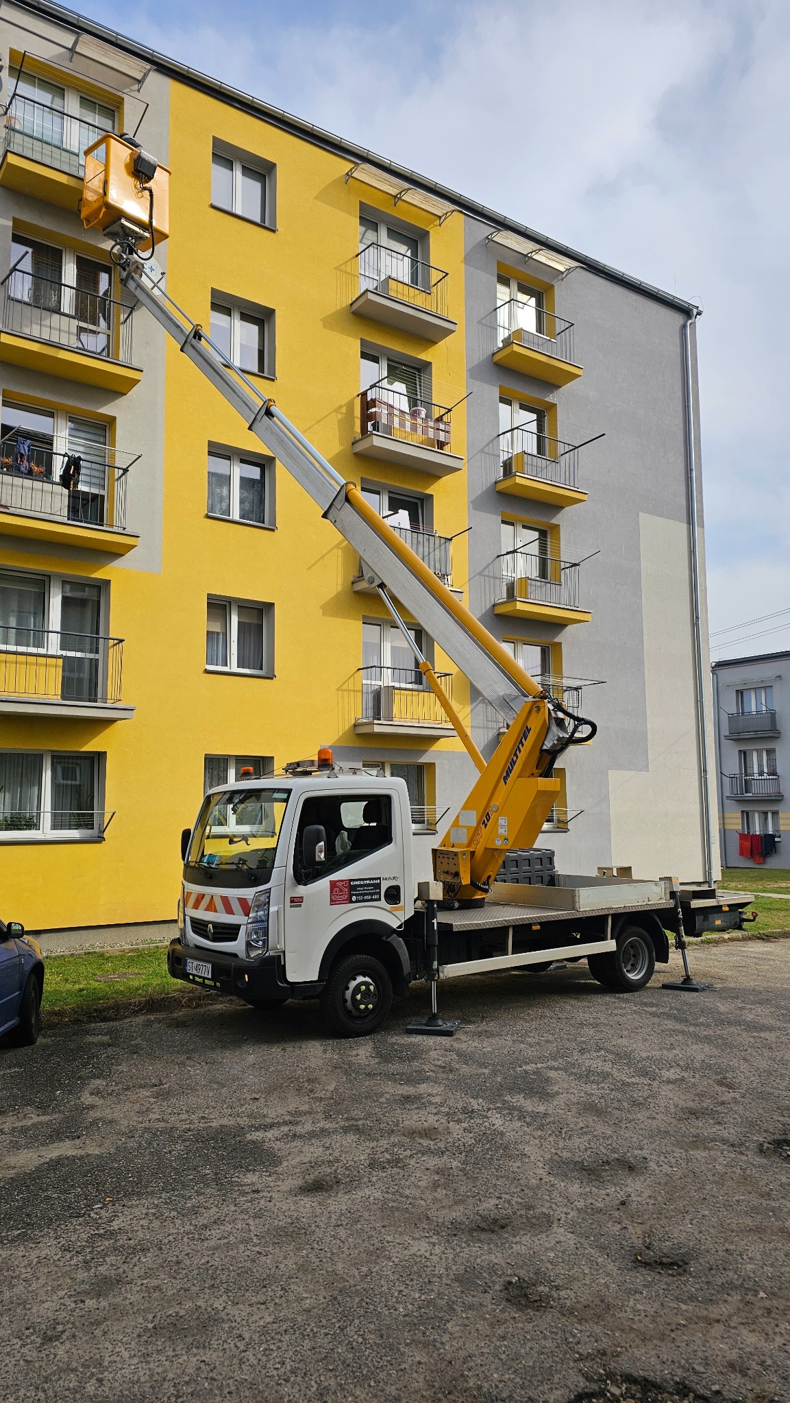 Żółty podnośnik koszowy z wysięgnikiem ustawiony przed blokiem mieszkalnym z żółtą i szarą elewacją, kosz na wysokości balkonu, widoczne detale konstrukcji i podpory stabilizujące pojazd.