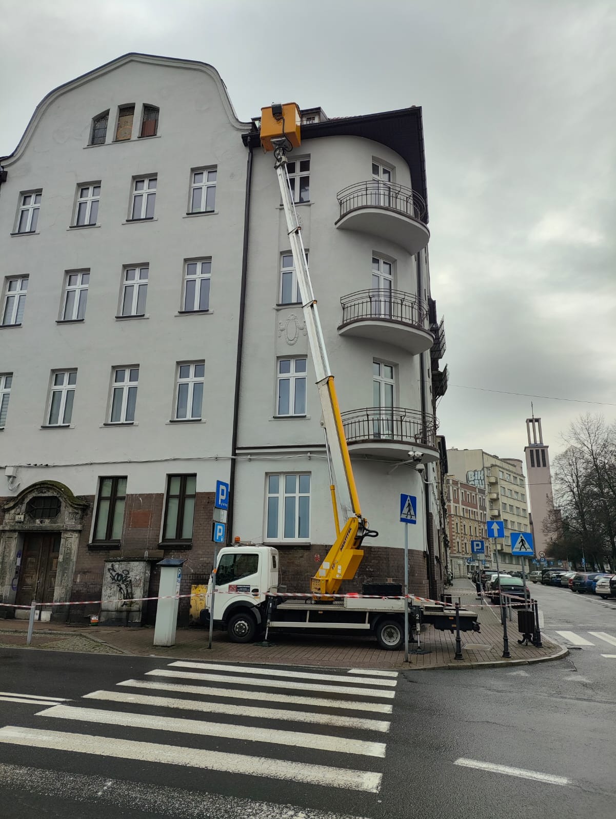Żółty podnośnik koszowy na białym samochodzie ustawiony przy narożnej kamienicy z balkonami, widok z poziomu ulicy na pasach, w tle kościół i zaparkowane samochody.