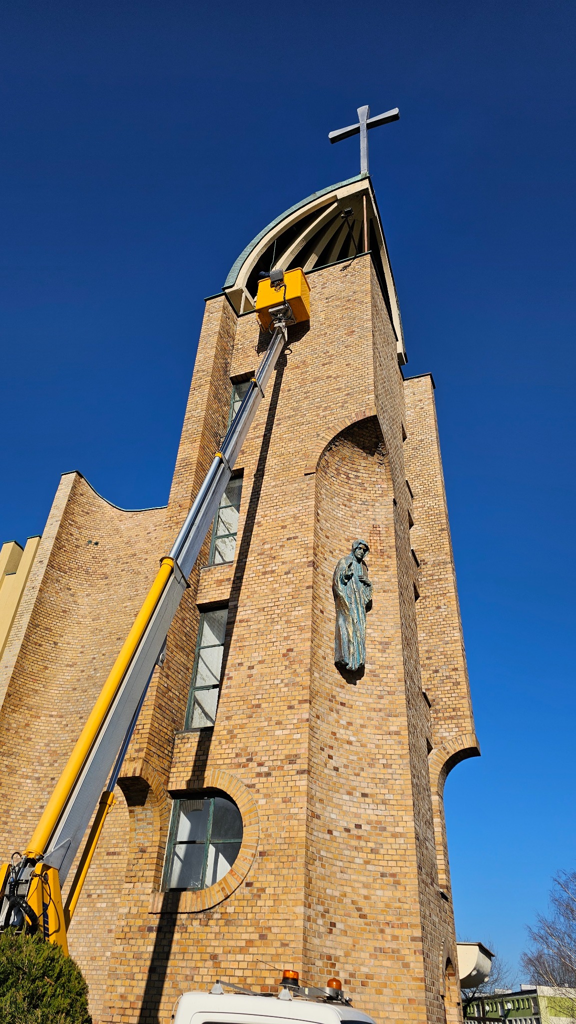 Żółty podnośnik koszowy sięga do ceglanej wieży kościoła z krzyżem na szczycie i posągiem świętego w niszy, na tle błękitnego nieba.