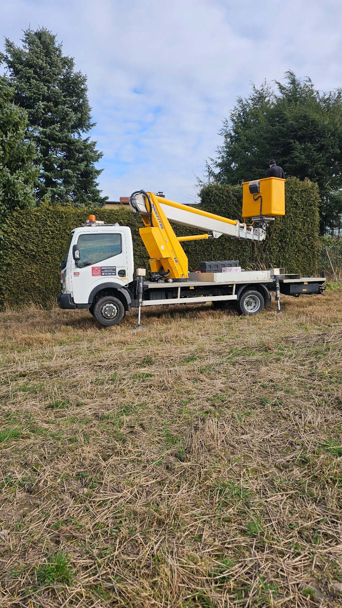 Biała ciężarówka z żółtym podnośnikiem koszowym, z pracownikiem w koszu, na tle wysokiego żywopłotu i drzew, na polu z uschniętą trawą.