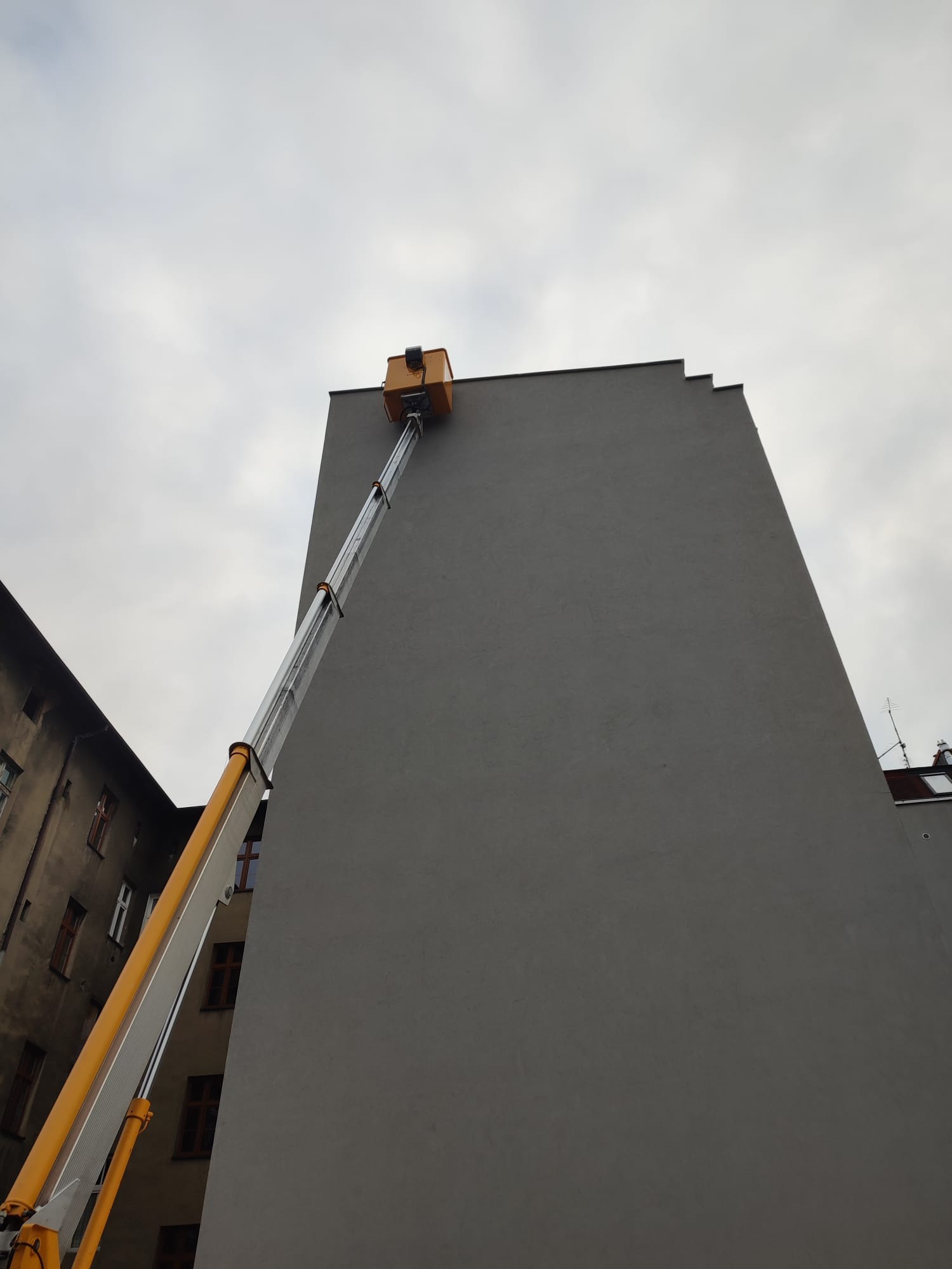 Żółto-srebrny podnośnik koszowy sięga do szczytu wysokiego, szarego budynku w pochmurny dzień, widok z dołu.