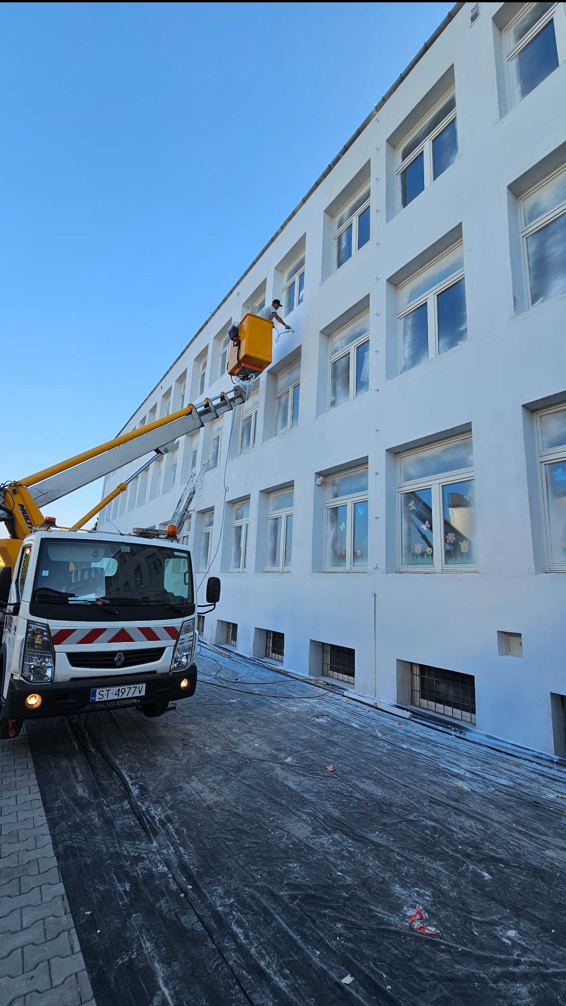 Pracownik w podnośniku koszowym, malujący jasną elewację budynku z licznymi oknami, widoczne zabezpieczenie podłoża folią, błękitne niebo w tle.