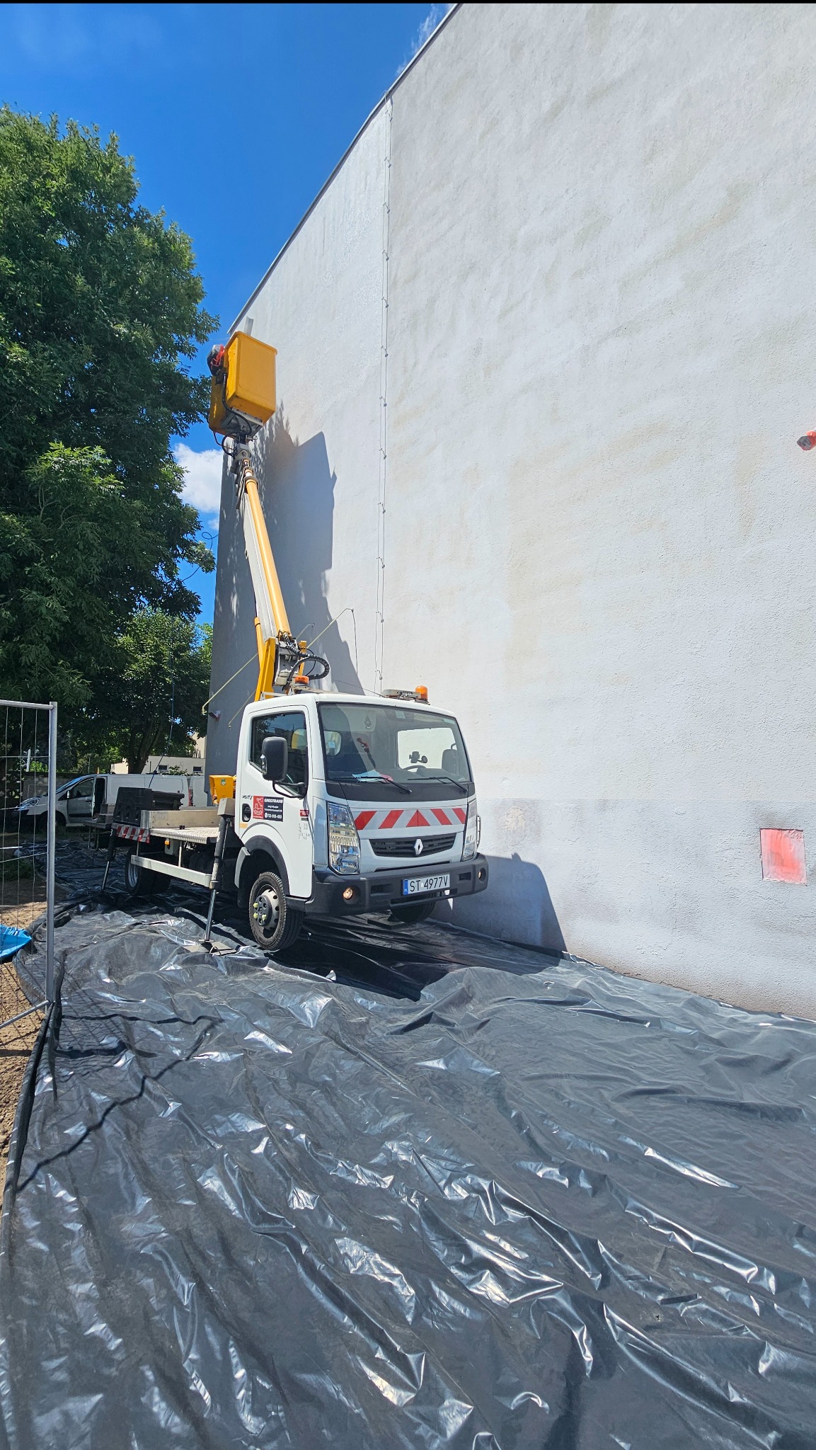 Żółty podnośnik koszowy przy białej ścianie budynku z zabezpieczeniem folią na ziemi.