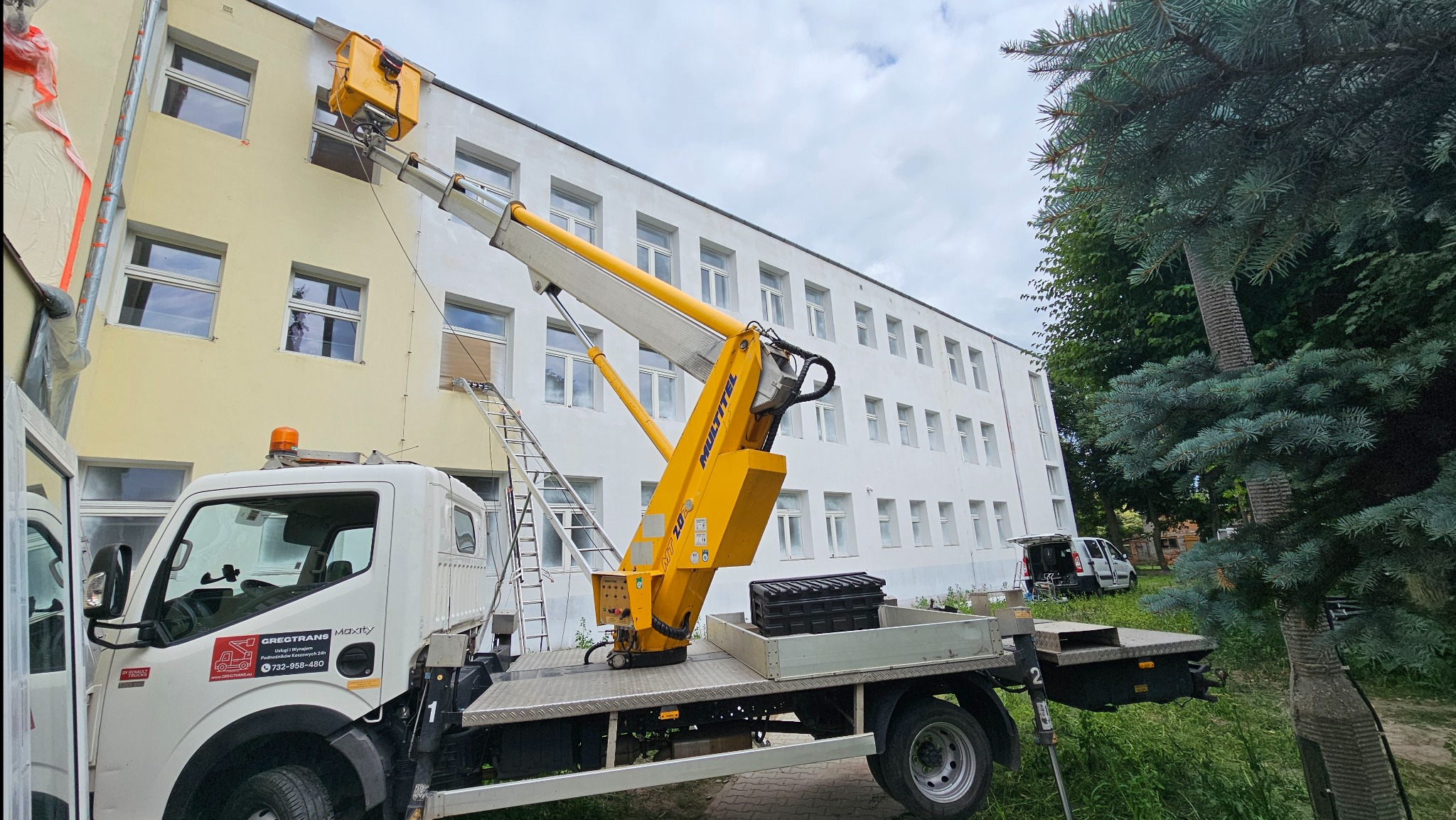 Żółty podnośnik koszowy na białej ciężarówce z logo firmy GREGTRANS, ustawiony przy dwukolorowym budynku z wieloma oknami, drabina oparta o ścianę, widoczne ślady prac na elewacji.