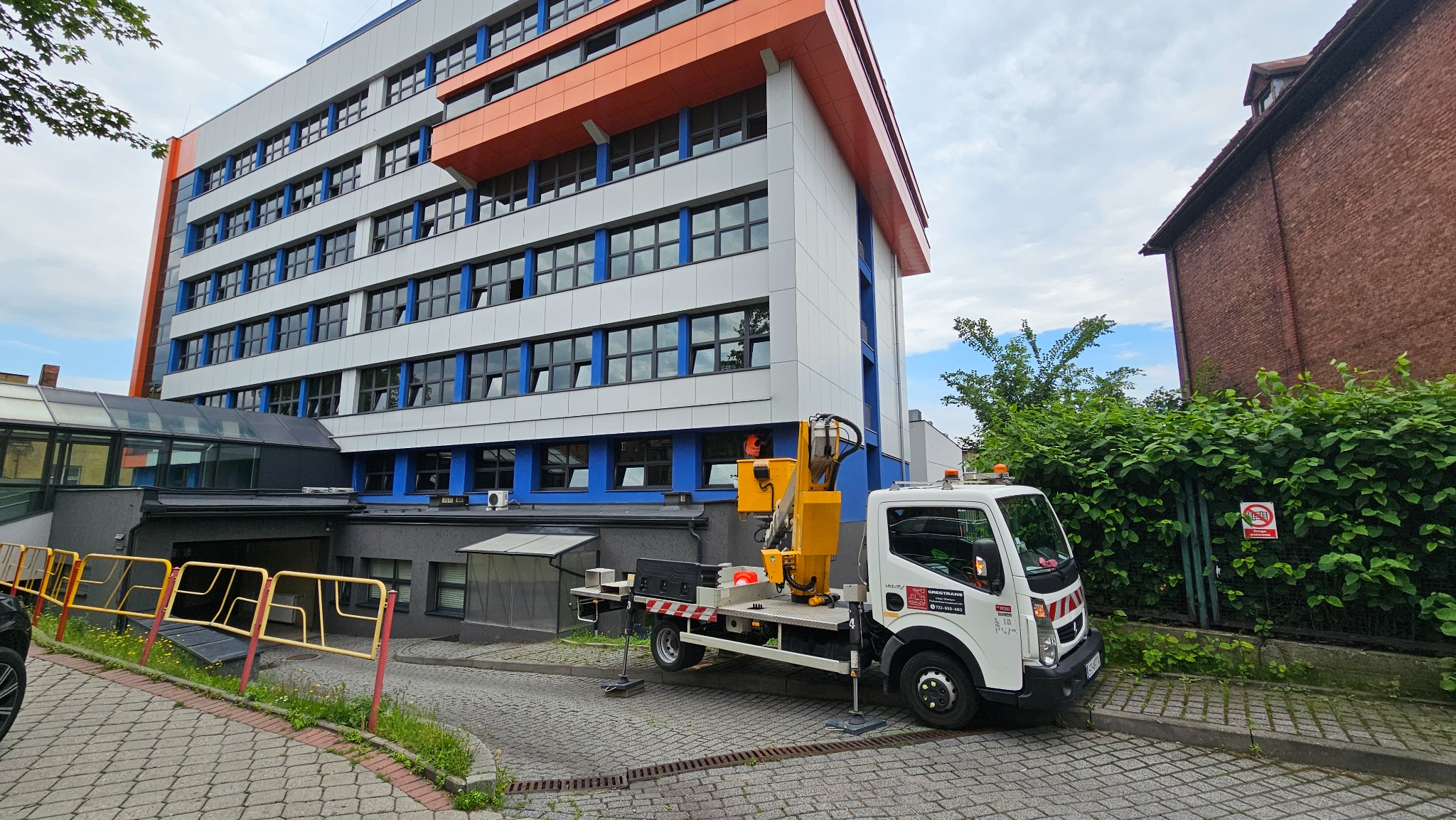 Żółty podnośnik koszowy na białej ciężarówce zaparkowany na brukowanej drodze przed nowoczesnym budynkiem biurowym z elementami pomarańczowymi i niebieskimi.