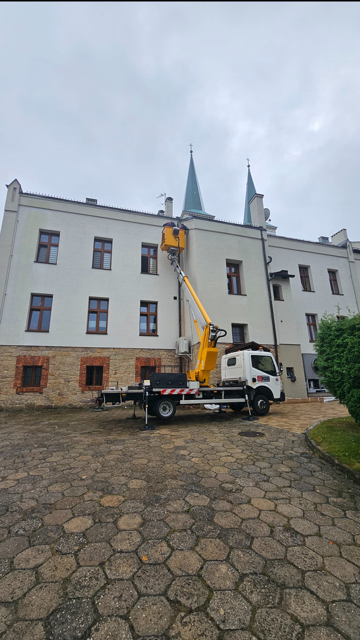 Żółty podnośnik koszowy na tle jasnej elewacji budynku z dwoma wieżami zwieńczonymi iglicami, pracownicy w koszu na wysokości okien.