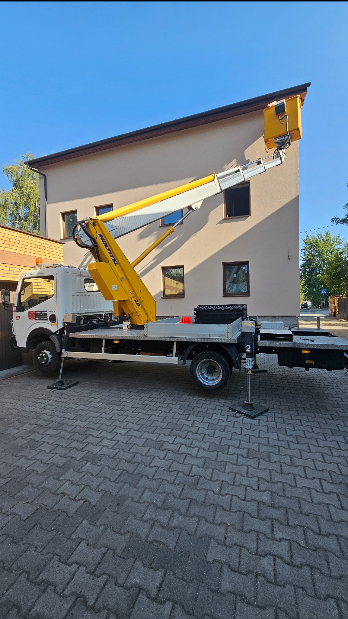 Żółto-biały podnośnik koszowy ustawiony na kostce brukowej przed dwupiętrowym budynkiem z beżową elewacją i brązowymi ramami okiennymi, z wysuniętym koszem w kierunku dachu.