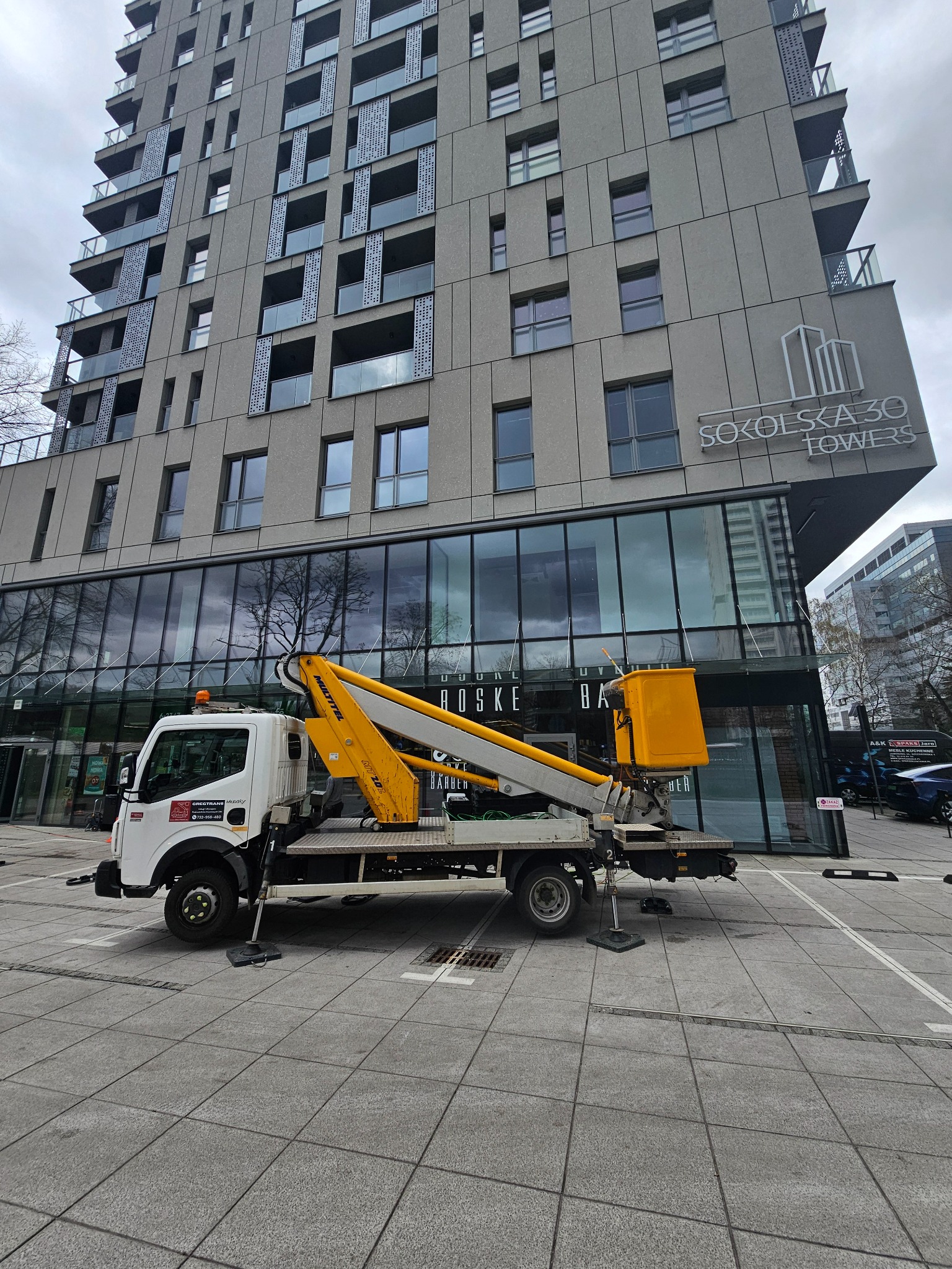 Żółty podnośnik koszowy na białej ciężarówce zaparkowany przed nowoczesnym budynkiem biurowym 'Sokolska 30 Towers' z dużą szklaną fasadą i szarymi panelami. Stabilizatory rozłożone na szarej kostce...