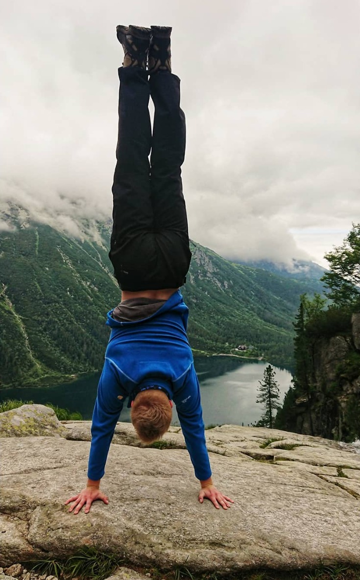 Mężczyzna w niebieskiej bluzie i ciemnych spodniach wykonuje stójkę na tle górskiego jeziora i zalesionych szczytów w pochmurny dzień.