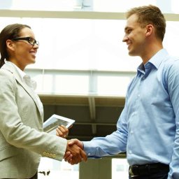 Marek Taborski - Uśmiechnięta kobieta w okularach i jasnym żakiecie, trzymająca tablet, wita się uściskiem dłoni z mężczyzną w błękitnej koszuli w jasnym, nowoczesnym wnętrzu.