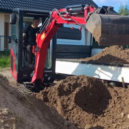 KOLTRA Monika Kaliszczak - Operator w czerwonej minikoparce Yanmar wysypuje ziemię do skrzyni ładunkowej białego samochodu dostawczego na tle domu z ciemnym dachem.