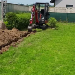 Czerwono-czarna mini-koparka Yanmar na trawniku obok wykopanego wąskiego rowu z brązową ziemią, w tle dom jednorodzinny i słup oświetleniowy.