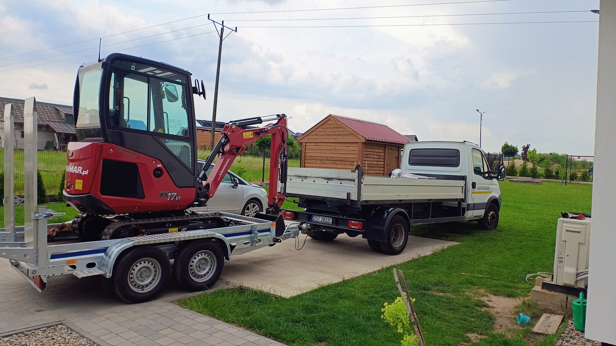 Czerwony mini-koparka Yanmar SV17VT na przyczepie, ciągnięta przez białą ciężarówkę Goritrans w Stargardzie; w tle domek narzędziowy i zabudowa jednorodzinna.