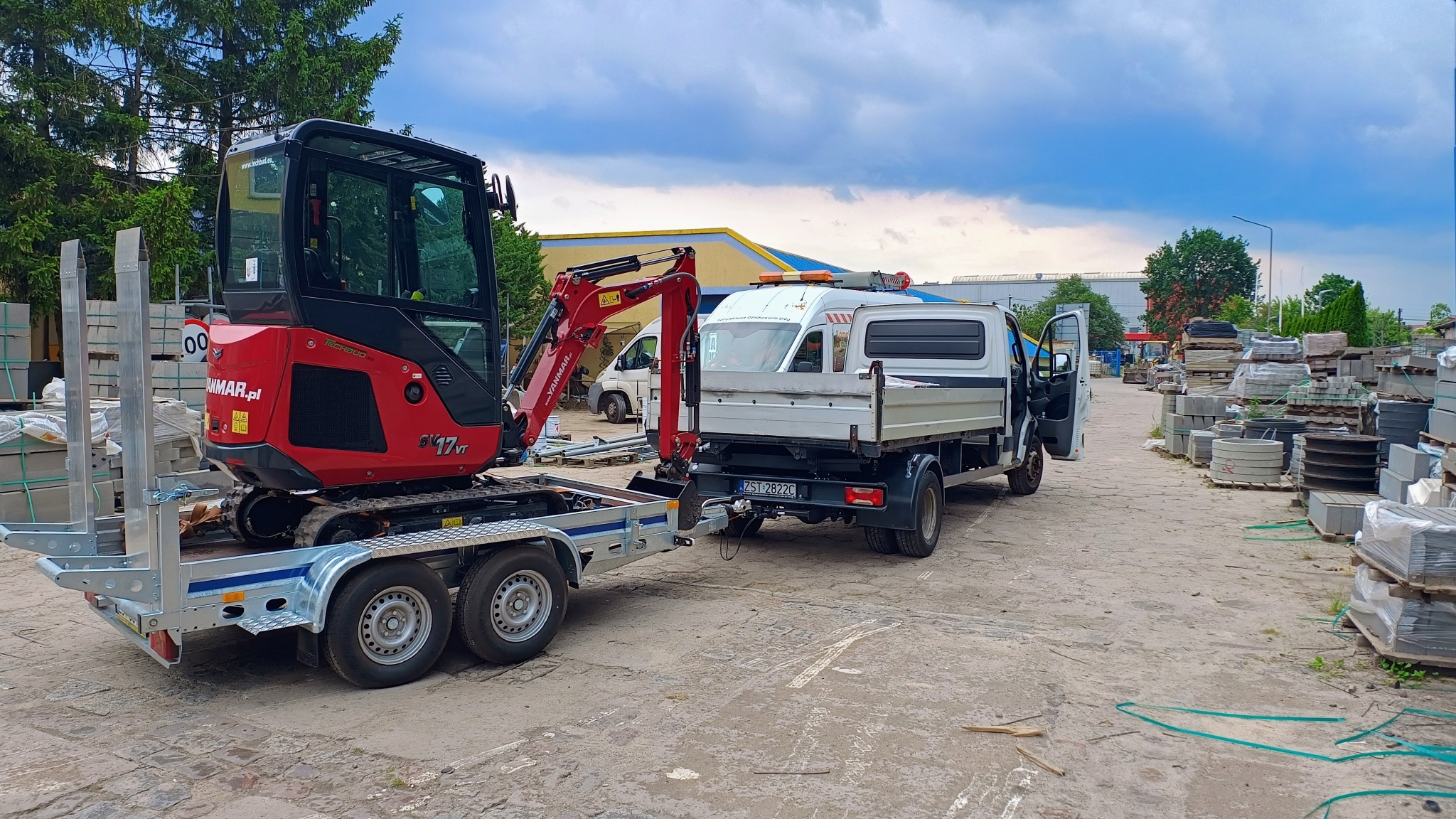 Czerwona mini koparka Yanmar SV17VT na przyczepie transportowej, podłączona do białego samochodu dostawczego z otwartymi drzwiami, w tle składowane materiały budowlane.