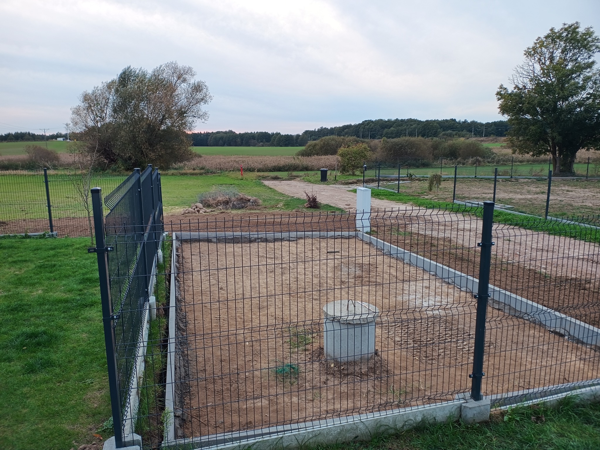 Wykonany drenaż na działce z betonowym zbiornikiem, zabezpieczony ogrodzeniem z siatki na tle zielonego krajobrazu.