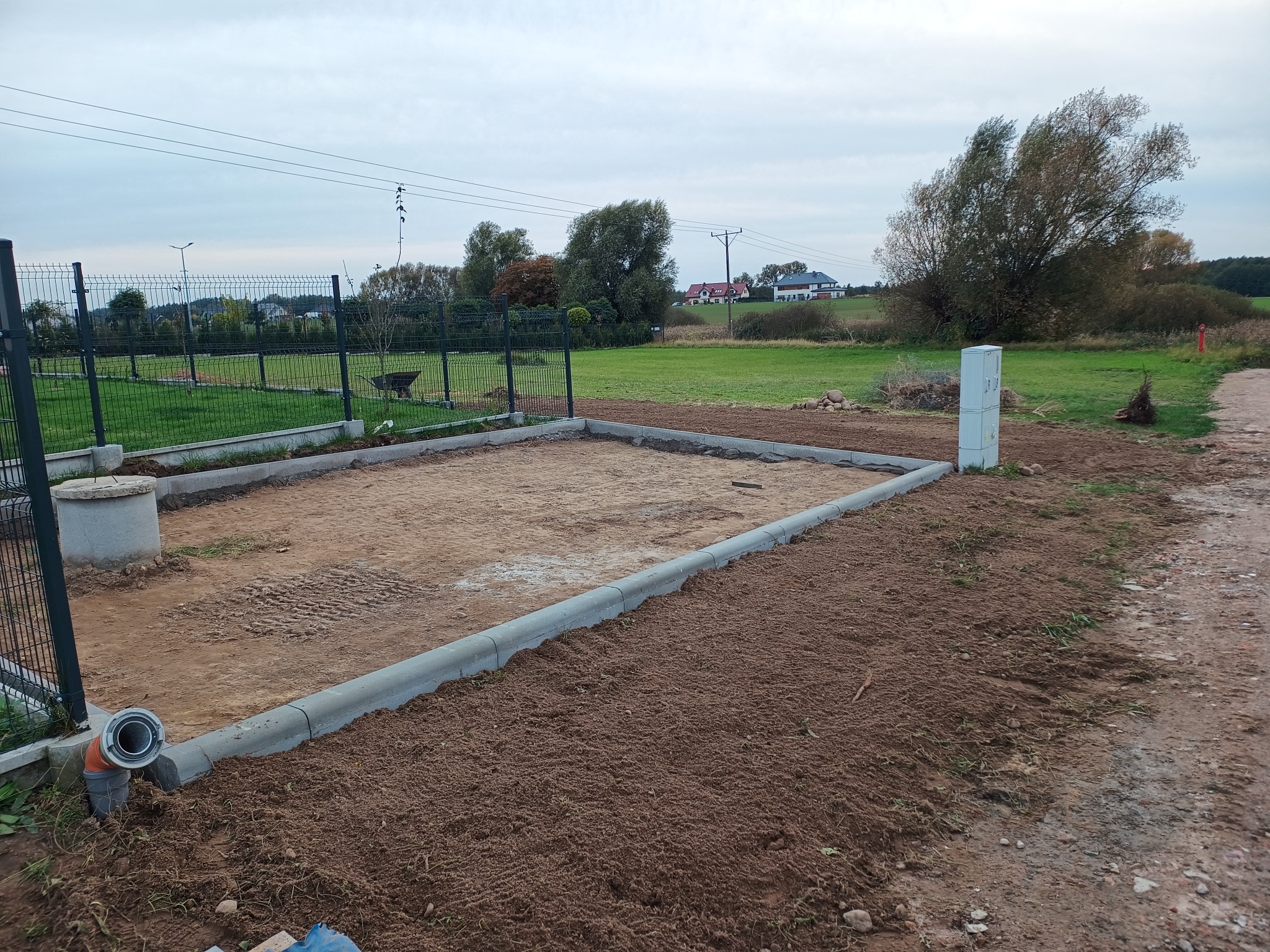 Przygotowany teren pod budowę z obrzeżami betonowymi, widoczny drenaż, ogrodzenie panelowe w tle oraz zabudowa jednorodzinna.