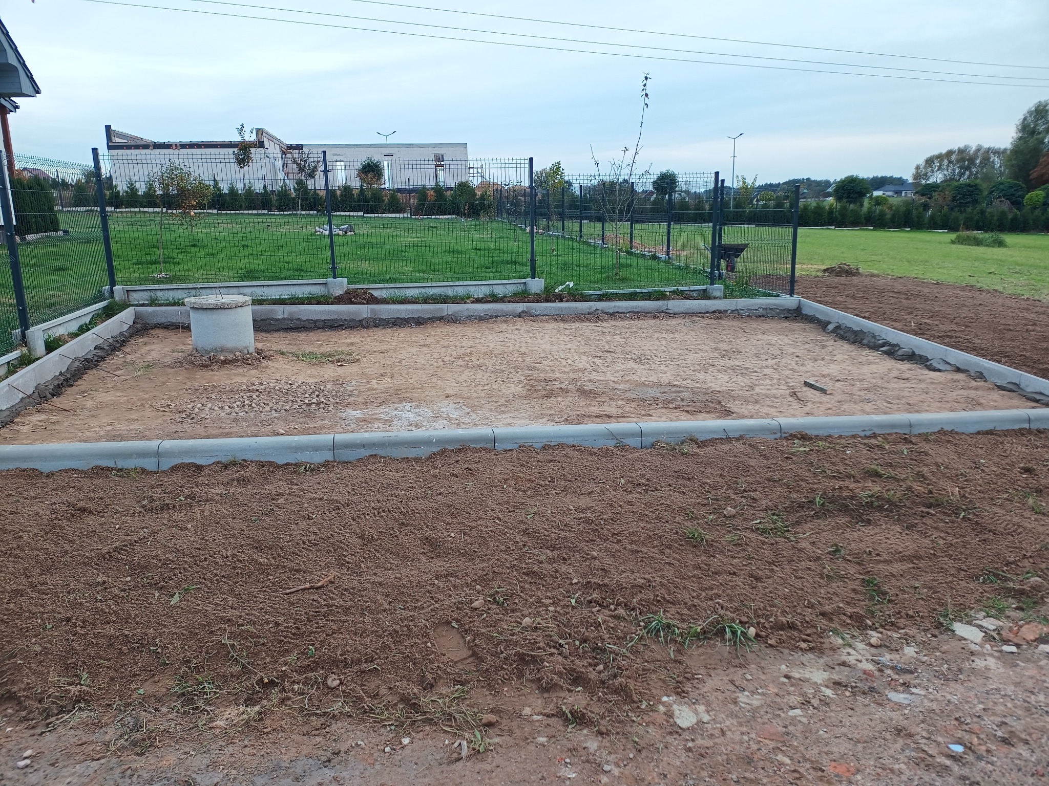 Przygotowany teren pod budowę z ułożonymi obrzeżami, widoczny betonowy krąg studzienki kanalizacyjnej i ogrodzenie posesji w tle.