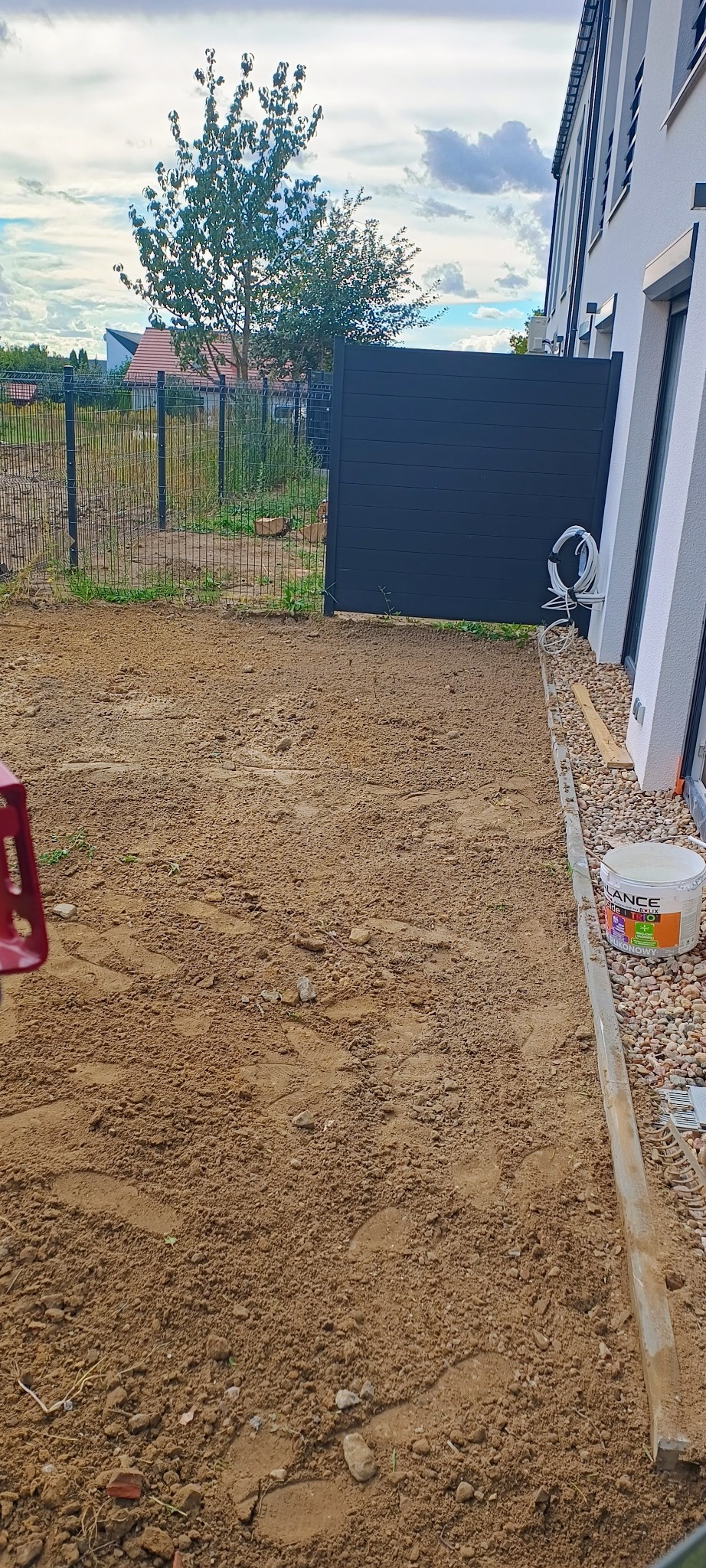 Przygotowany teren pod budowę, widoczne ślady stóp na ziemi, obrzeża z kostki, wiadro z klejem budowlanym i zwinięty kabel przy ścianie budynku.