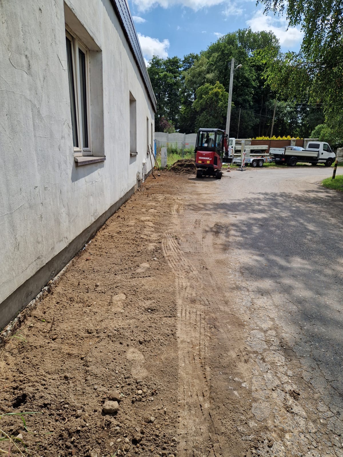 Czerwony minikoparka Yanmar stoi obok budynku z przygotowanym pasem ziemi, prawdopodobnie pod fundamenty, z widocznymi śladami opon i zaparkowanym samochodem dostawczym w tle.