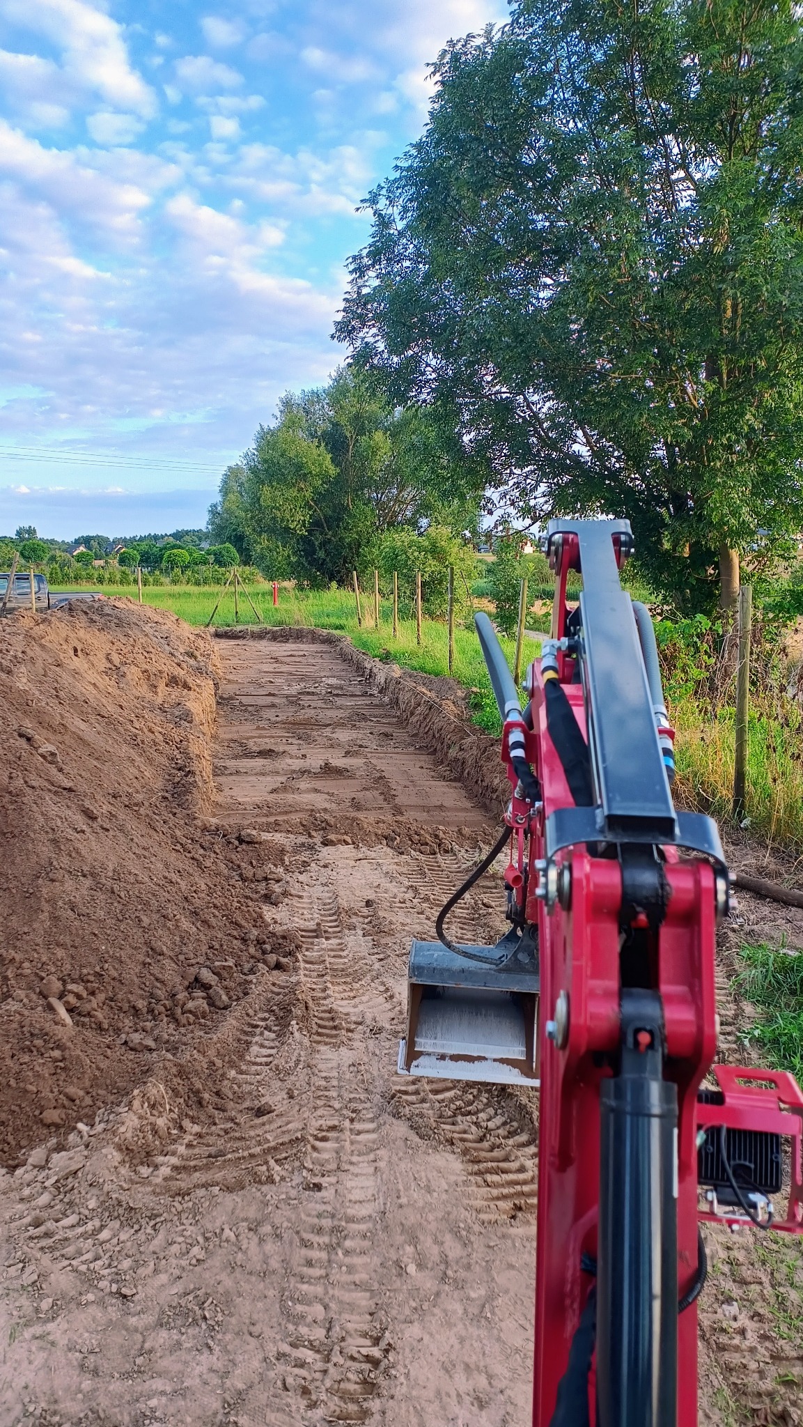 Czerwona mini koparka na placu budowy, wykop pod fundament z widocznymi śladami gąsienic, świeżo usypana ziemia i zieleń w tle.