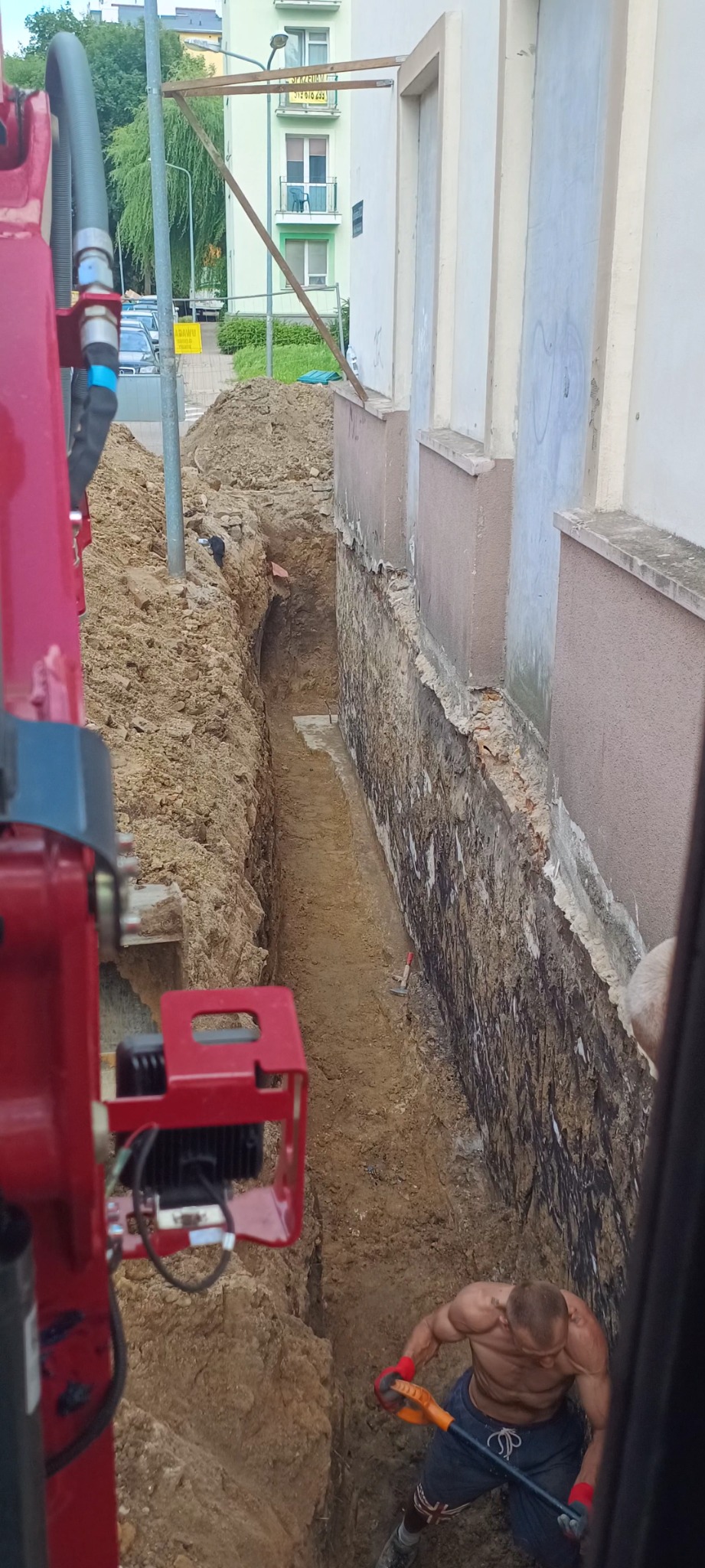 Głęboki wykop wąskoprzestrzenny przy ścianie budynku, widok z kabiny koparki, muskularny mężczyzna kopie szpadlem na dnie wykopu.