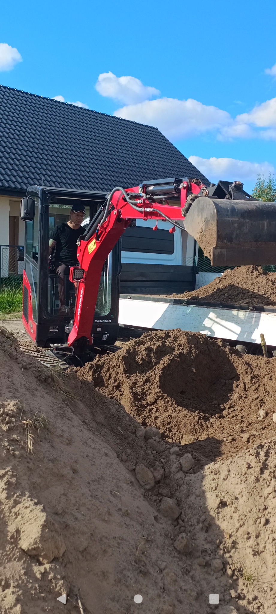 Operator w czerwonej minikoparce Yanmar wysypuje ziemię do skrzyni ładunkowej białego samochodu dostawczego na tle domu z ciemnym dachem.