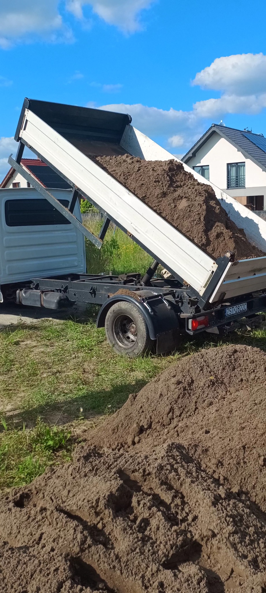 Ciężarówka z otwartą skrzynią wysypuje ziemię na trawnik przed nowym domem z panelami słonecznymi na dachu.