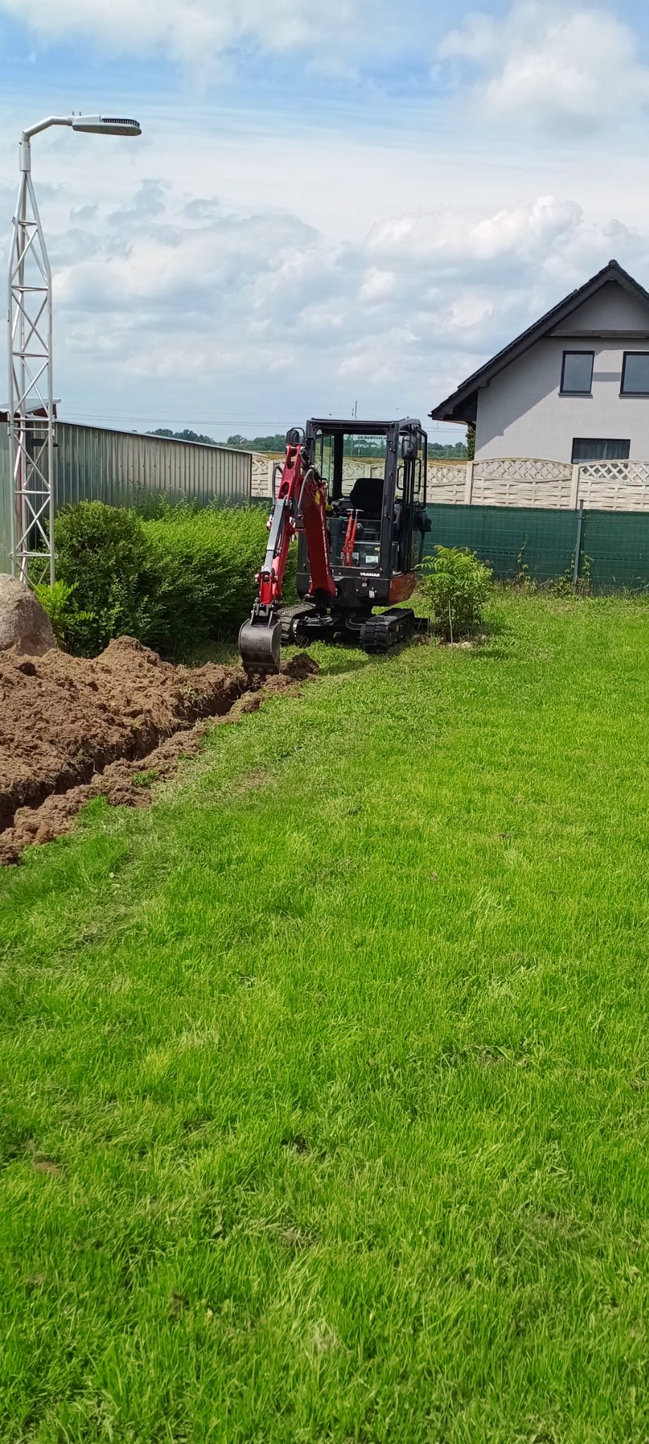 Czerwono-czarna mini-koparka Yanmar na trawniku obok wykopanego wąskiego rowu z brązową ziemią, w tle dom jednorodzinny i słup oświetleniowy.
