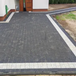 Ułożona, ciemna kostka brukowa z białymi pasami obramowania, tworząca ścieżkę i podjazd przed budynkiem. W tle widoczny trawnik i krzewy.