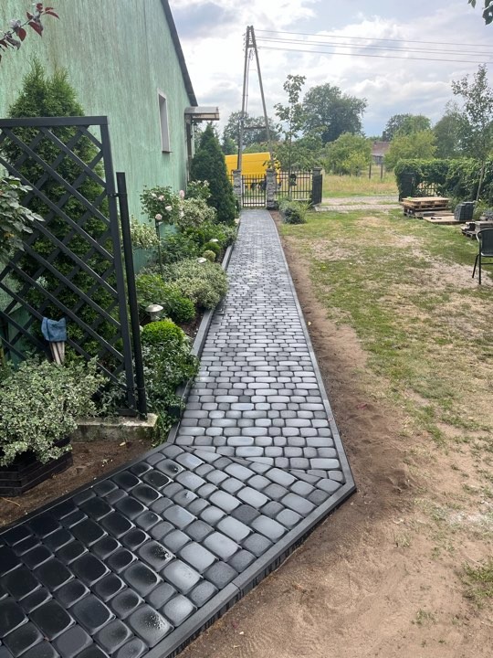 Ścieżka z szarej kostki brukowej prowadząca od ciemnej, mokrej kostki wzdłuż zielonego domu z ozdobną kratą i roślinami, zakończona bramą w tle.