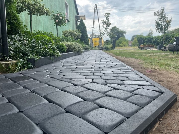 Ułożona ścieżka z grafitowej kostki brukowej o nieregularnym kształcie, zakończona obrzeżem, prowadząca w kierunku metalowej bramy na tle zieleni i pochmurnego nieba.