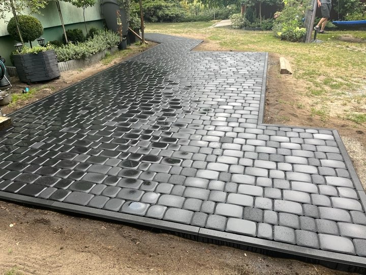 Świeżo ułożona, czarna kostka brukowa w kształcie litery L, obramowana ciemnoszarą krawędzią, tworząca ścieżkę w ogrodzie z widocznymi roślinami w tle.