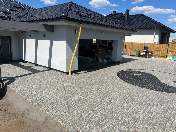 Podjazd z kostki granitowej ułożonej we wzór koła przed garażem domu jednorodzinnego z czarnym dachem i panelami słonecznymi, widoczne kosze na śmieci i bambusowe ogrodzenie w tle.