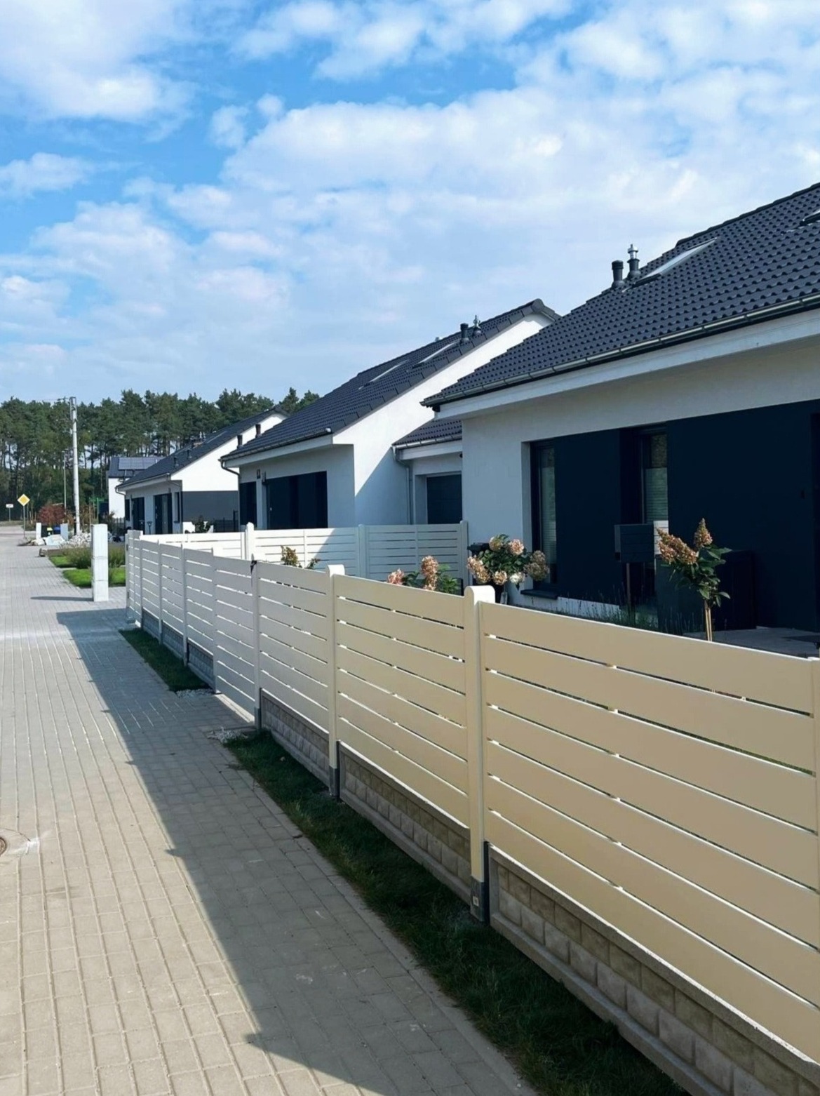 Nowoczesne, poziome, białe ogrodzenie panelowe z podmurówką wzdłuż chodnika i szeregu domów jednorodzinnych z ciemnymi oknami i dachami w słoneczny dzień.