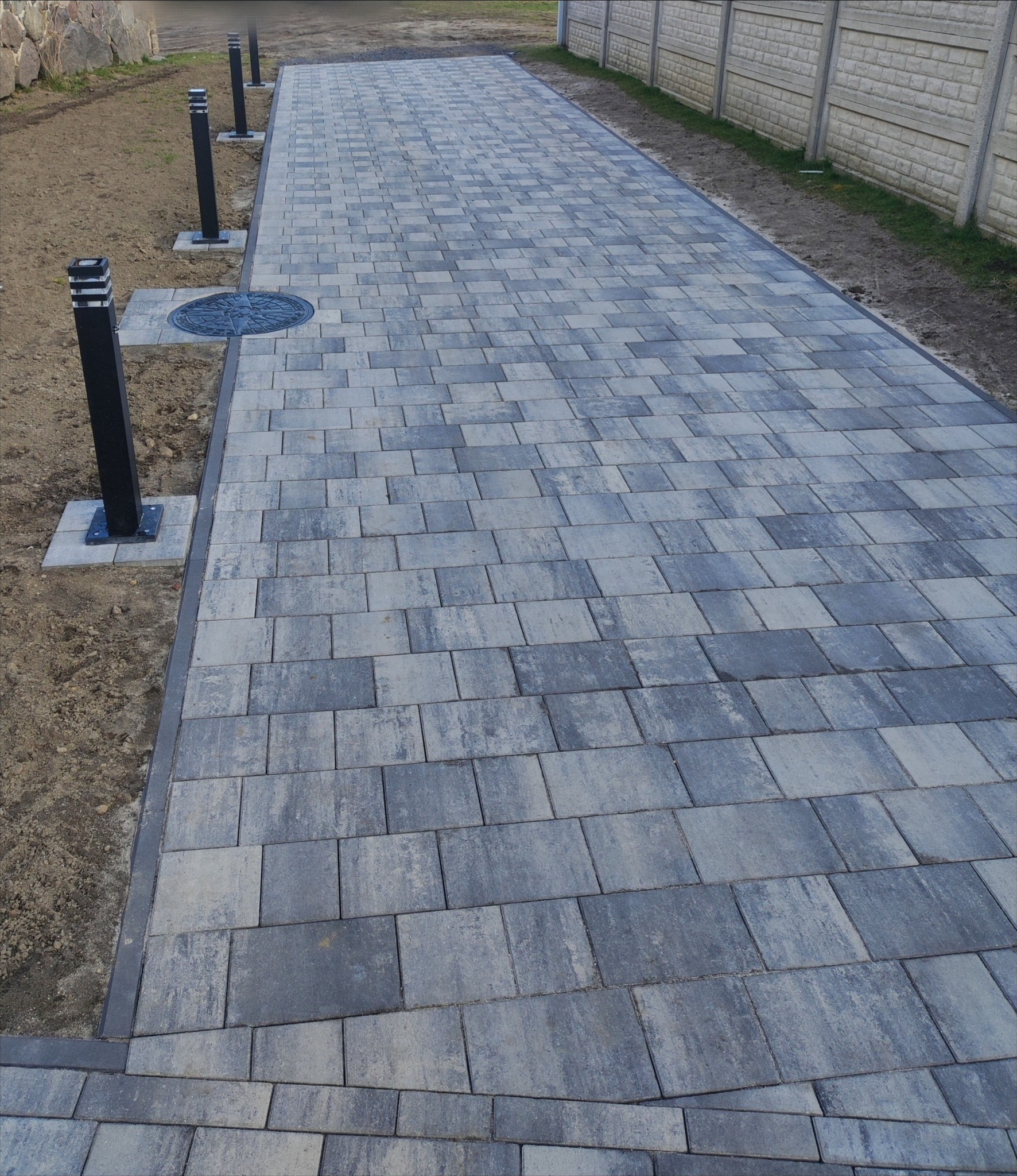 Ułożona szara kostka brukowa tworząca ścieżkę z oświetleniem i betonowym ogrodzeniem w tle. Widoczny właz kanalizacyjny.