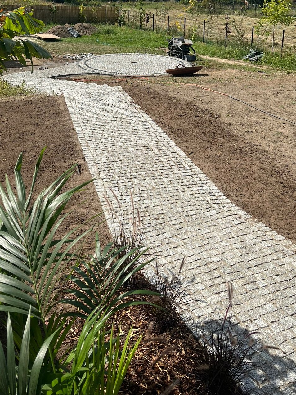 Granitowa ścieżka wijąca się przez ogród, z roślinami na pierwszym planie i ubijarką w tle, sugerująca świeżo zakończone prace brukarskie.