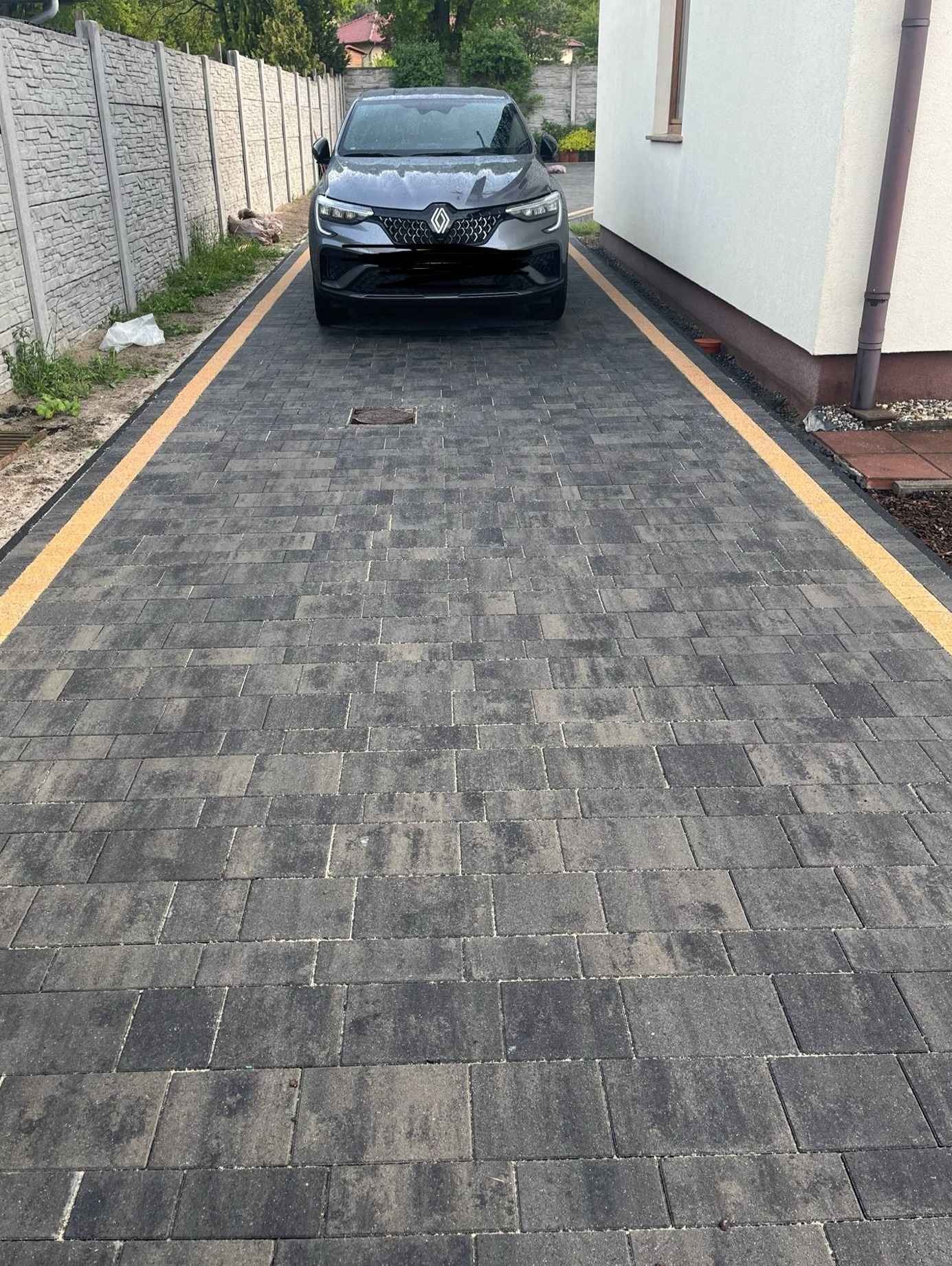 Ułożona kostka brukowa w odcieniach szarości z żółtymi pasami po bokach, na której stoi czarny samochód. W tle biały budynek i szary betonowy płot.