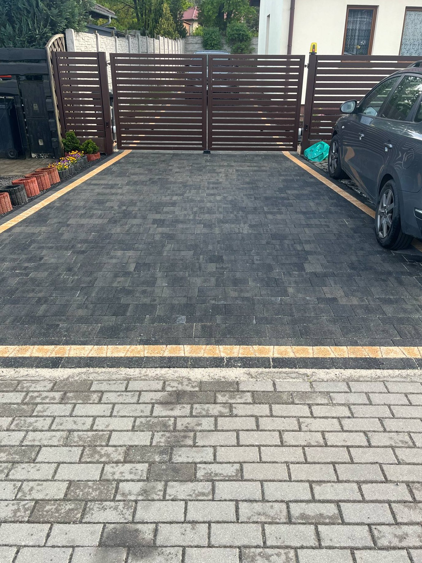 Nowoczesny podjazd z ciemnej kostki brukowej z żółtymi akcentami, zakończony bramą panelową. Widoczny fragment auta. Ułożona kostka przed domem.