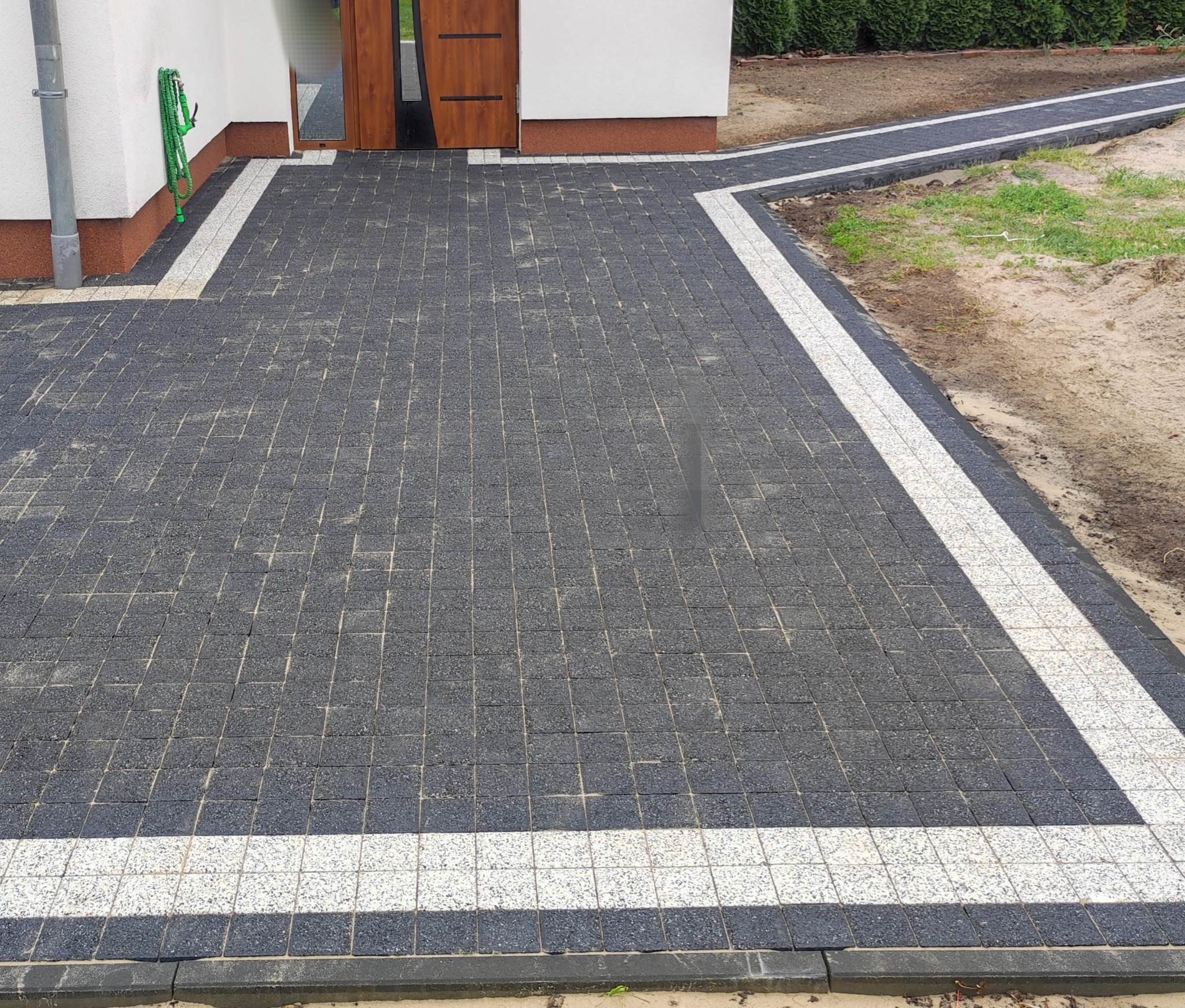 Ułożona, ciemna kostka brukowa z białymi pasami obramowania, tworząca ścieżkę i podjazd przed budynkiem. W tle widoczny trawnik i krzewy.
